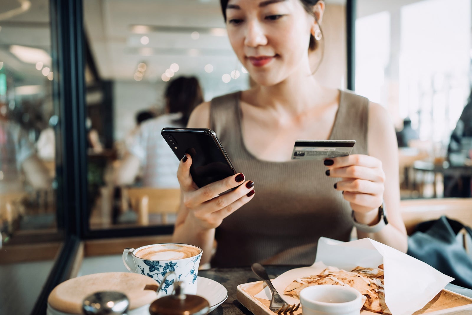 Beautiful smiling young Asian woman managing online banking with smartphone and making mobile payment with credit card on hand while having meal in a restaurant