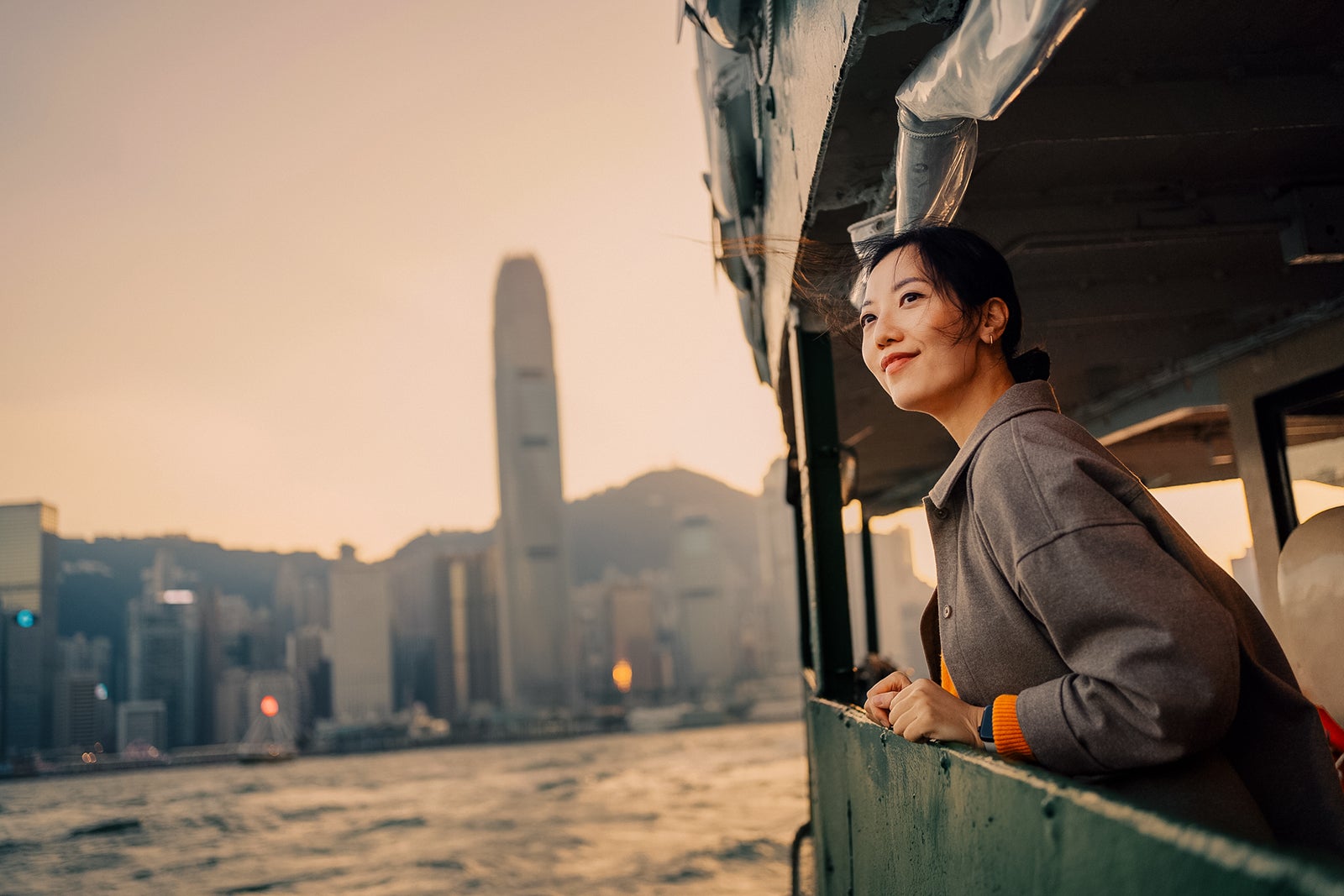 Smiling young Asian female traveller enjoying the spectacular iconic city skyline of Hong Kong and Victoria harbour while riding on the Star Ferry at dusk. Exploring local cultures in the city. Travel and vacation concept