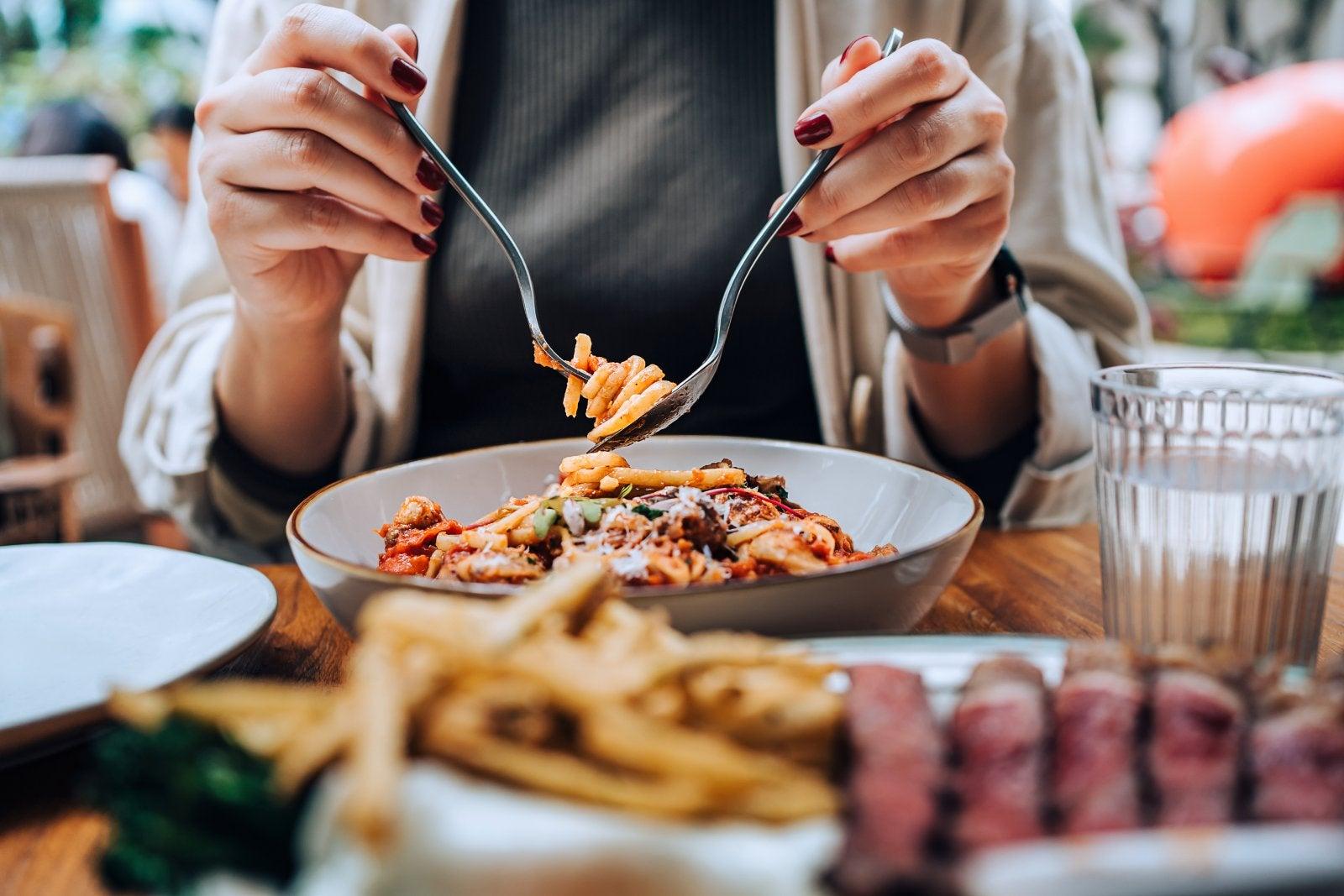Close-up, mid-section of young Asian woman sitting at dining table holding cutleries, twirling freshly served spaghetti bolognese and enjoying her meal in restaurant. Dining out. People, food and lifestyle